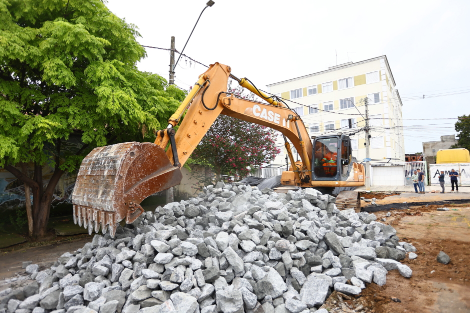 Escavadeira trabalha preenchendo o buraco com brita. Engenheiros aparecem ao fundo.