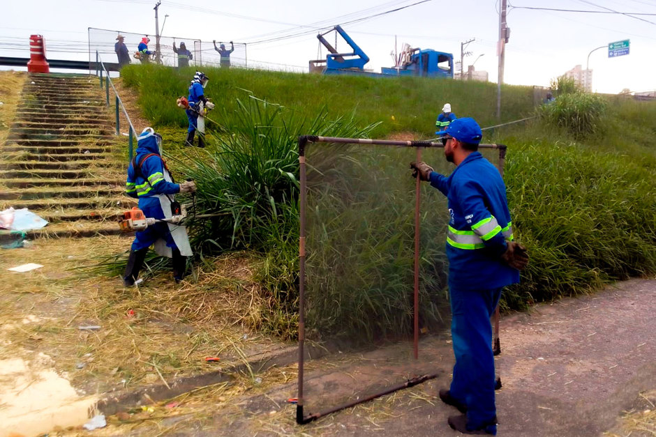 Equipe de roçada (em uniformes azuis) realiza serviço em via pública
