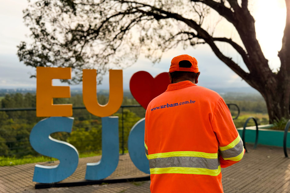Trabalhador da Urbam aparece varrendo a calçada do banhado, ao fundo o sol se põe e destaca o letreiro escrito "Eu amo SJC"