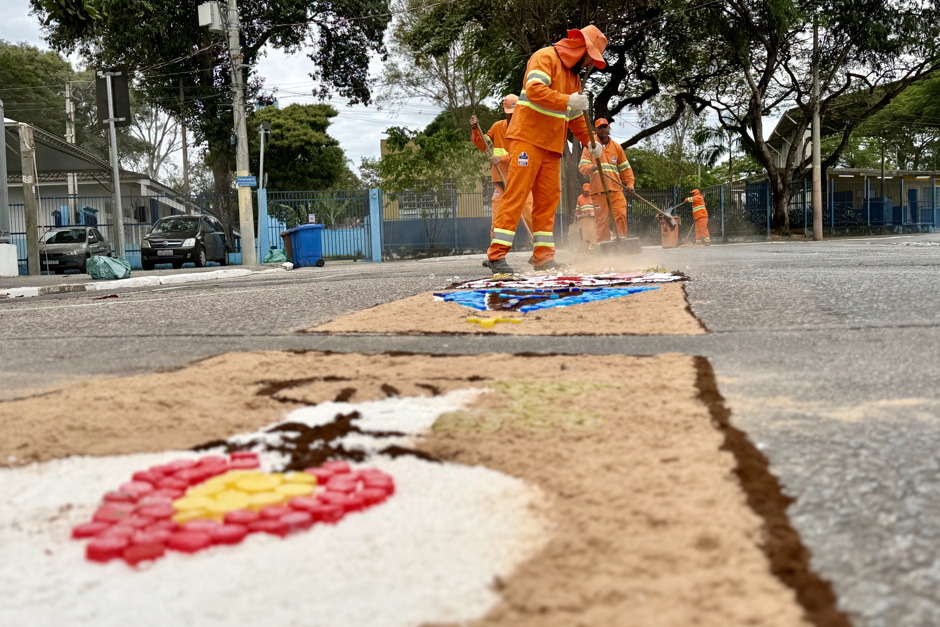 Tradicional tapete de Corpus Christi aparece em destaque enquanto agentes ambientais da Urbam, vestidos de uniformes laranjas, varrem o chão ao fundo da imagem