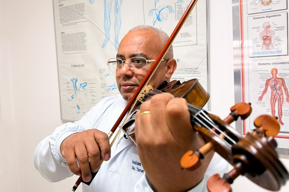 Isaías Hilário toca o seu violino vestido com seu jaleco de tanatopraxista